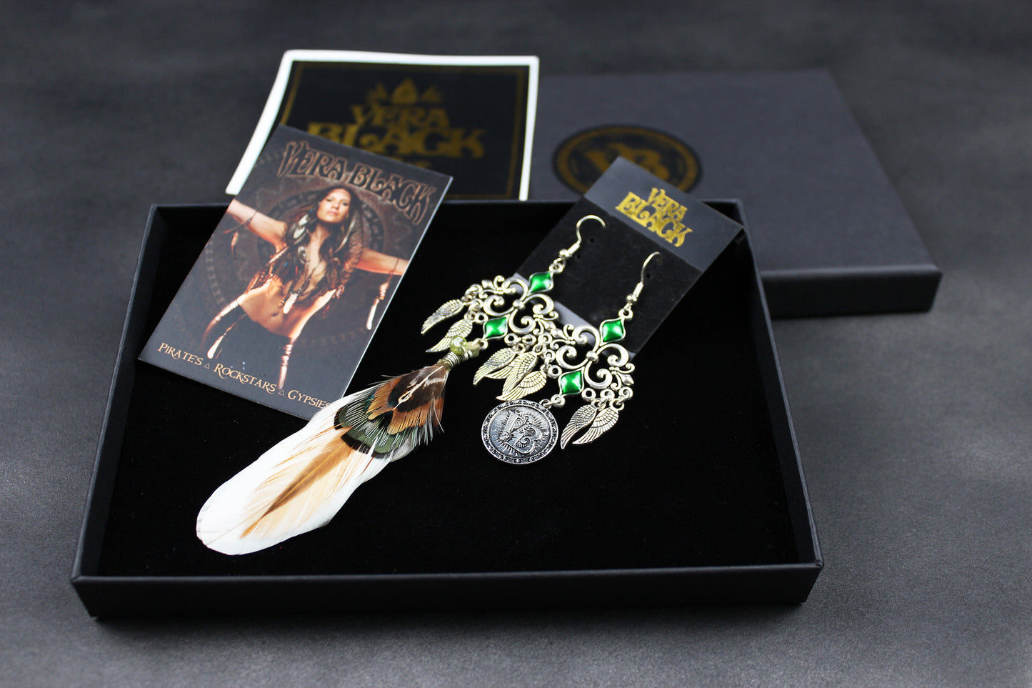 Pair of earrings with feathers and green stones in a black box with packaging on a dark background