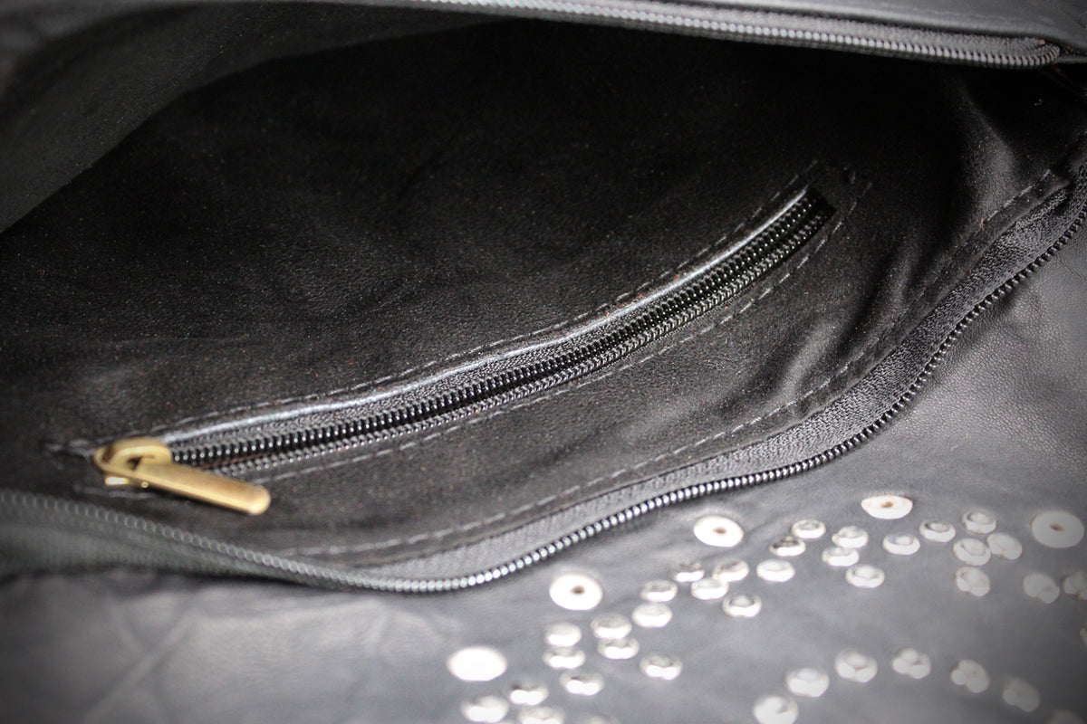 Close-up of a black leather or faux-leather bag interior showing a zippered pocket with a gold-tone zipper pull and scattered silver studs on the exterior surface.