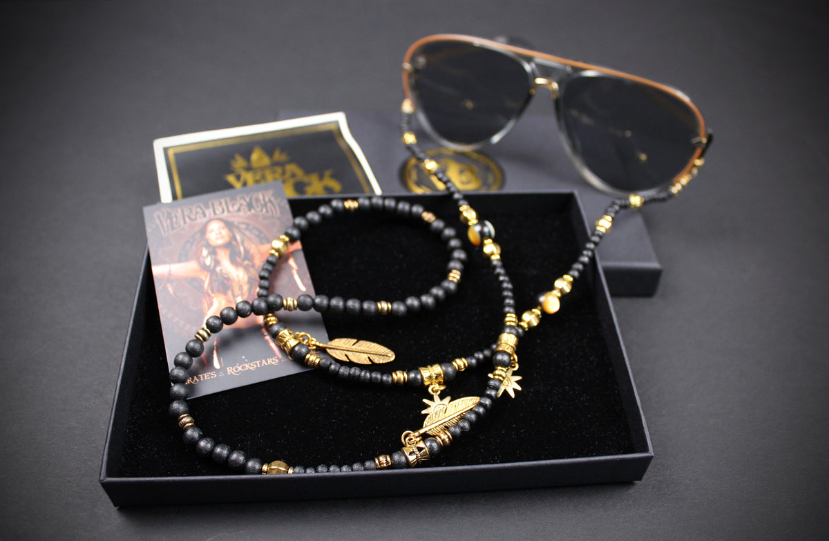A black beaded necklace with gold-toned charms and spacer beads, displayed in a black presentation box alongside sunglasses, a card, and a small square box on a dark background.