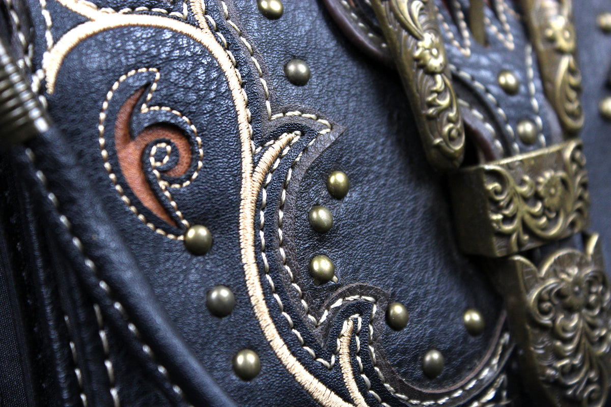 Close-up of a western-style leather bag or belt featuring dark navy leather, white contrast stitching, brass studs, and ornate antique bronze metal hardware.