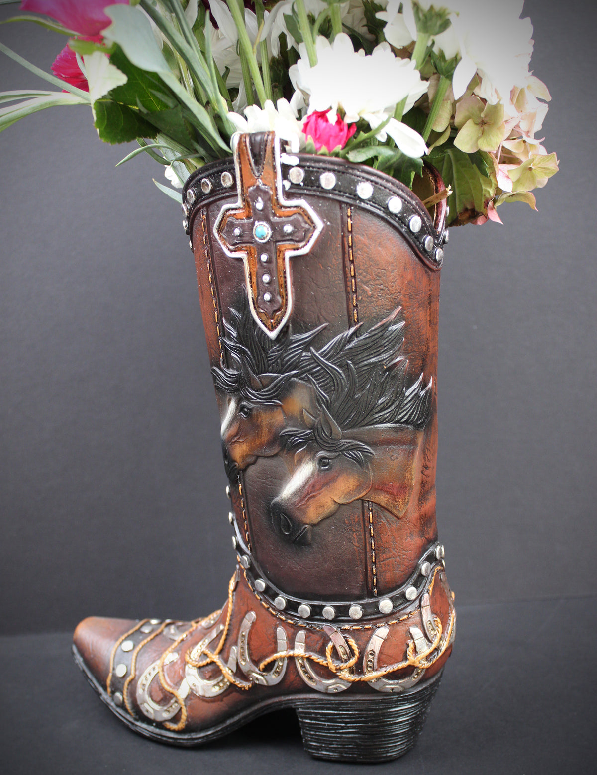 A brown leather cowboy boot vase filled with white and pink flowers, featuring hand-tooled western designs including a horse head, a cross, and silver and gold accents, set against a dark gray background.