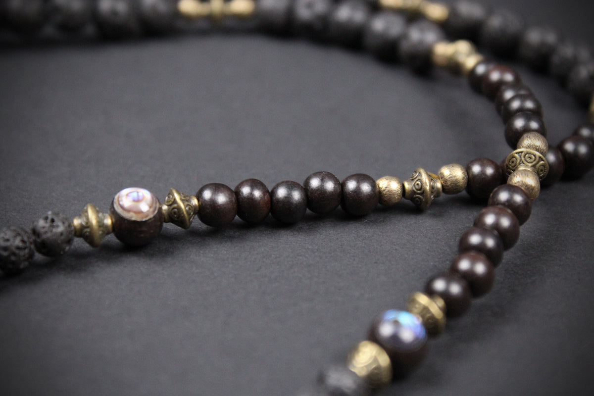 Dark brown wooden prayer beads with antique bronze metal spacers and a central iridescent pearl bead, displayed on a dark textured background.