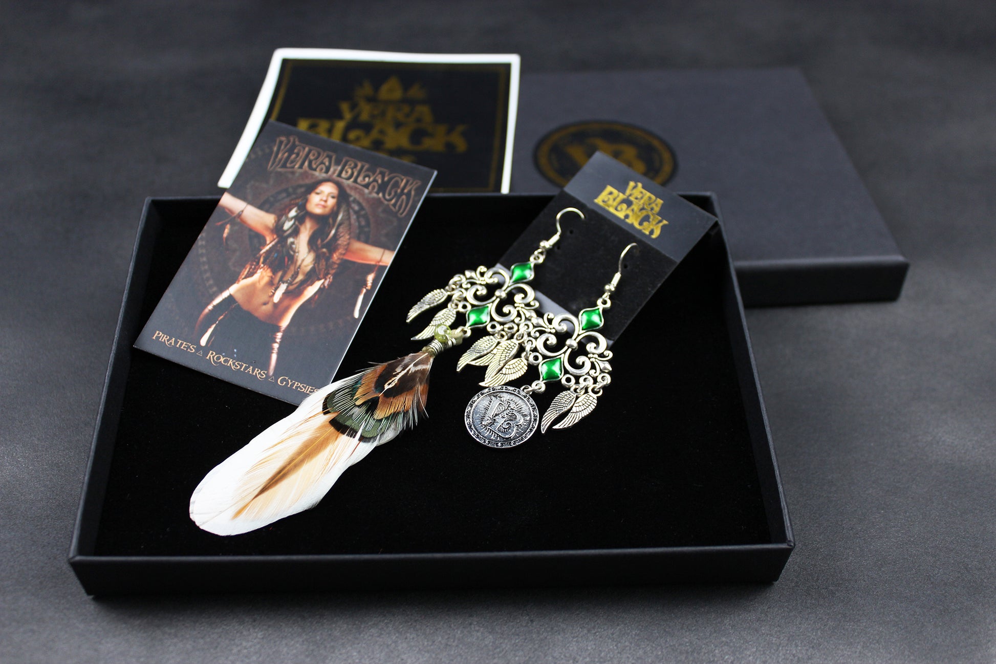 Pair of earrings with feathers and green stones in a black box with packaging on a dark background