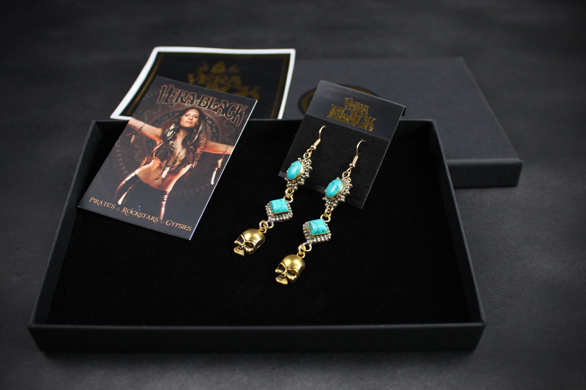 Gold and turquoise earrings in a black box with branded cards on a dark background