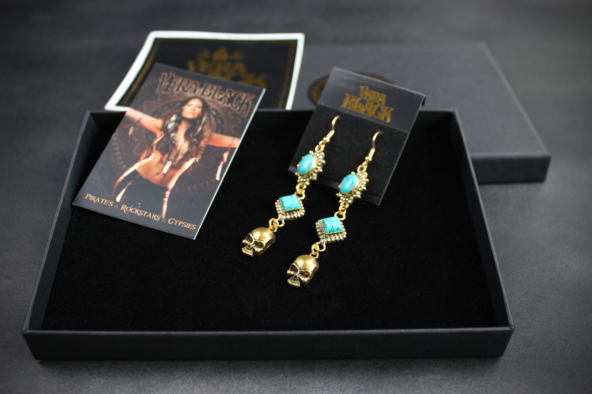 Gold earrings with turquoise stones on a black display box with a card featuring a woman.