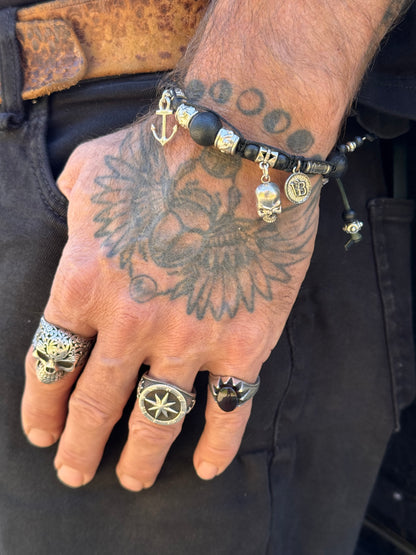 A close-up of a tattooed hand wearing multiple silver-toned rings and a beaded bracelet with skull and moon charms, paired with black pants and a brown belt.