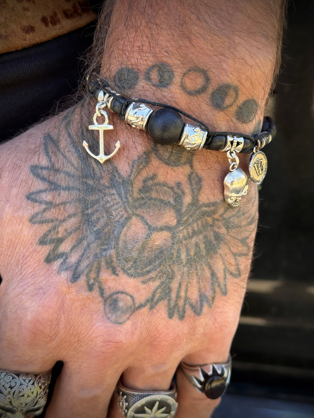A close-up of a tattooed hand wearing a black beaded bracelet with silver-toned charms, including an anchor, skull, and moon phases, along with silver rings.