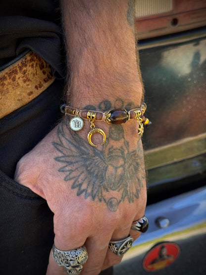 A close-up of a tattooed hand wearing a beaded charm bracelet with gold-toned rings and a crescent moon charm, set against a blurred outdoor background.