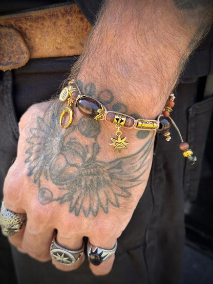 A close-up of a tattooed hand wearing a beaded bracelet with gold-toned charms, including a sun and moon pendant, alongside silver rings.