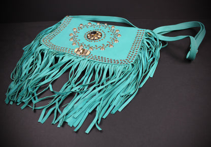 A turquoise fringe shoulder bag with gold-tone studs and a central medallion embellishment, displayed on a dark background.