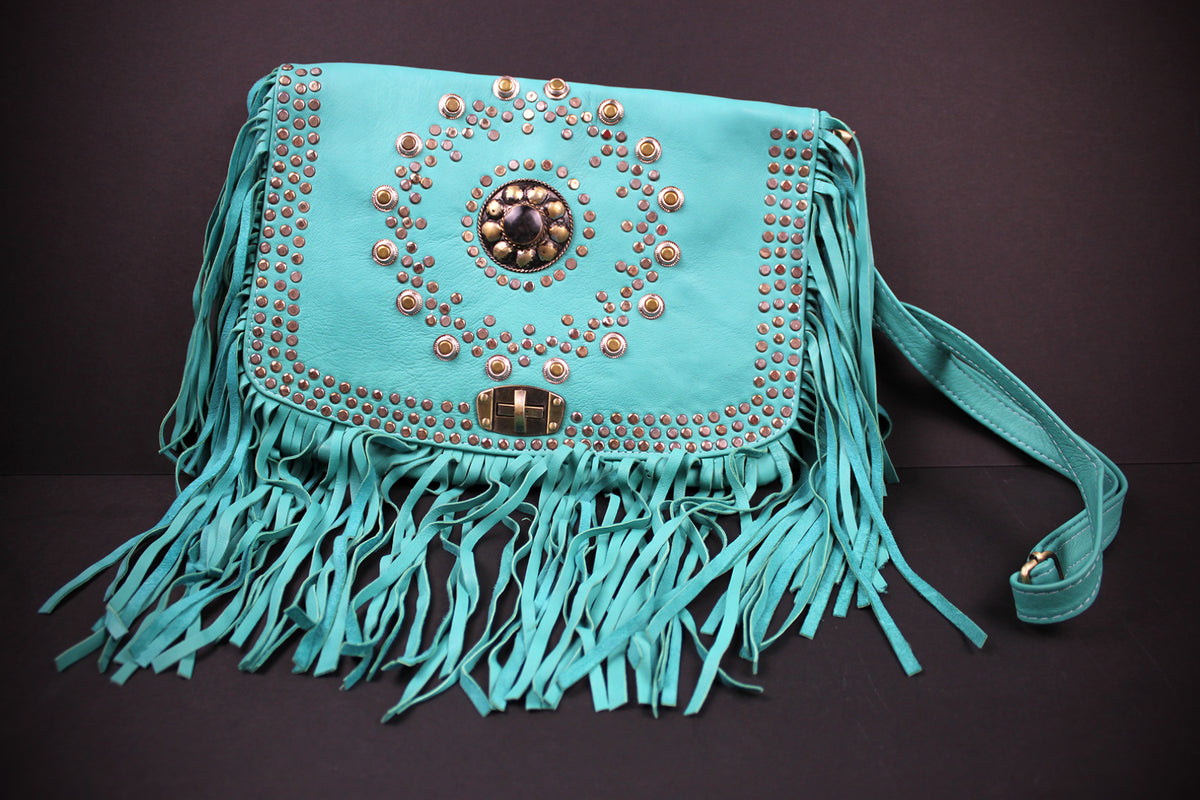 A turquoise blue leather shoulder bag with long fringe, studded details, and a central rhinestone embellishment, shown against a dark background.