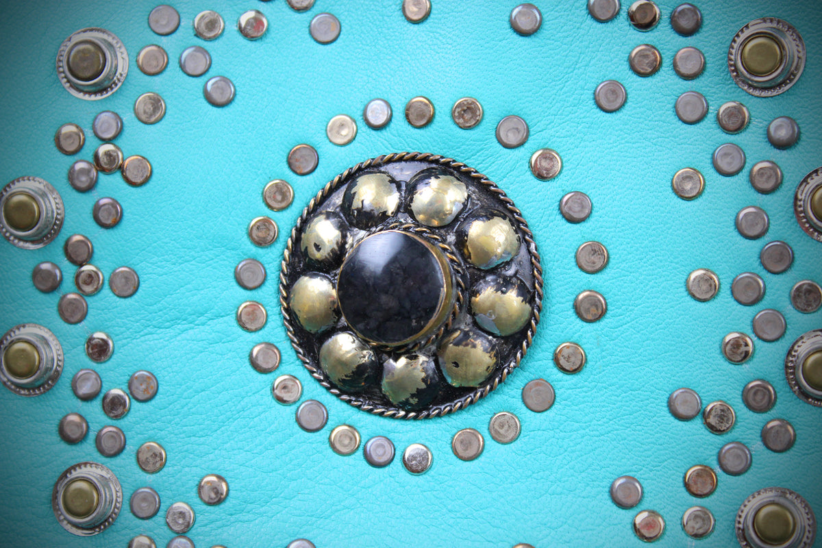 A close-up of turquoise leather adorned with numerous silver-toned studs and a large central decorative medallion featuring a dark center and metallic accents.