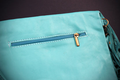 A close-up of a light turquoise blue faux leather bag featuring a horizontal zippered pocket with a gold-tone pull tab and matching blue zipper track, set against a dark background.