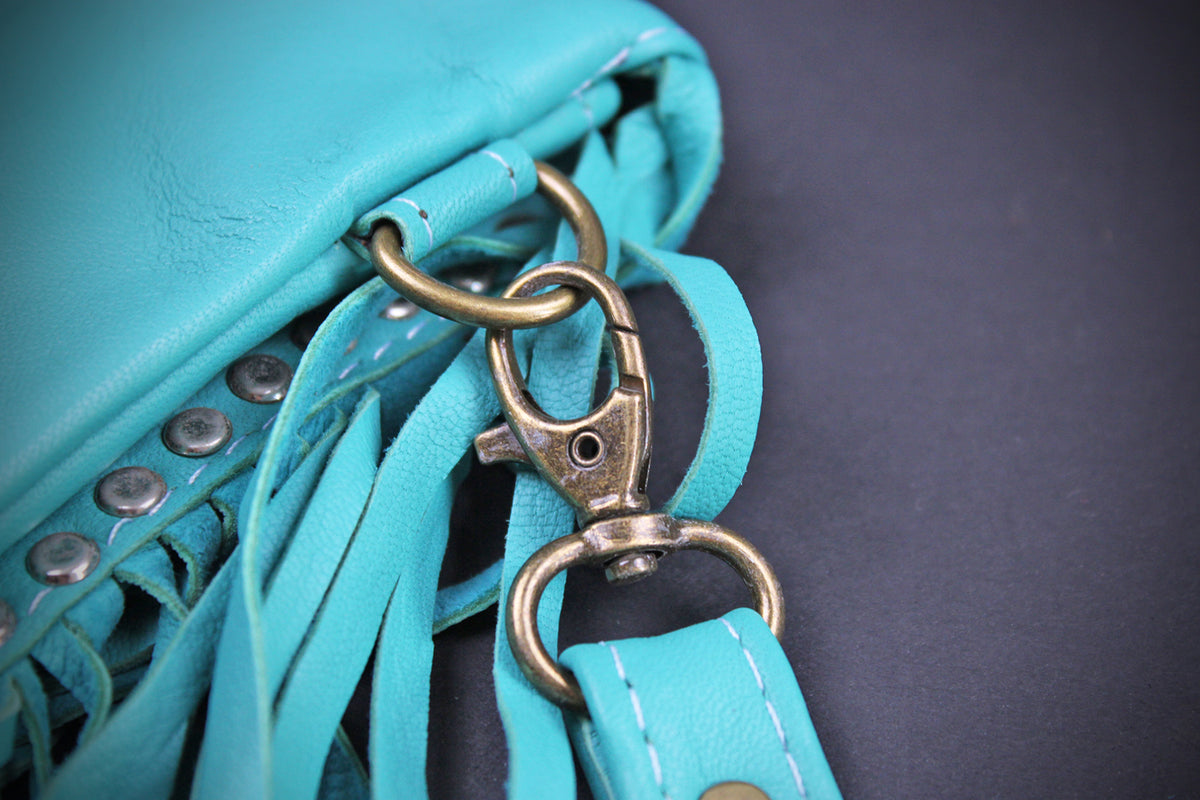 A close-up of a turquoise blue leather fringe bag with antique bronze metal hardware, including a lobster clasp and D-ring, on a dark gray background.