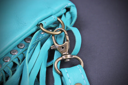 A close-up of a turquoise blue leather fringe bag with antique bronze metal hardware, including a lobster clasp and D-ring, on a dark gray background.
