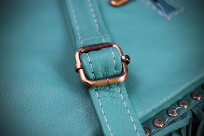Close-up of a teal leather strap with white contrast stitching and a distressed bronze buckle, attached to a matching teal bag with visible metal studs.