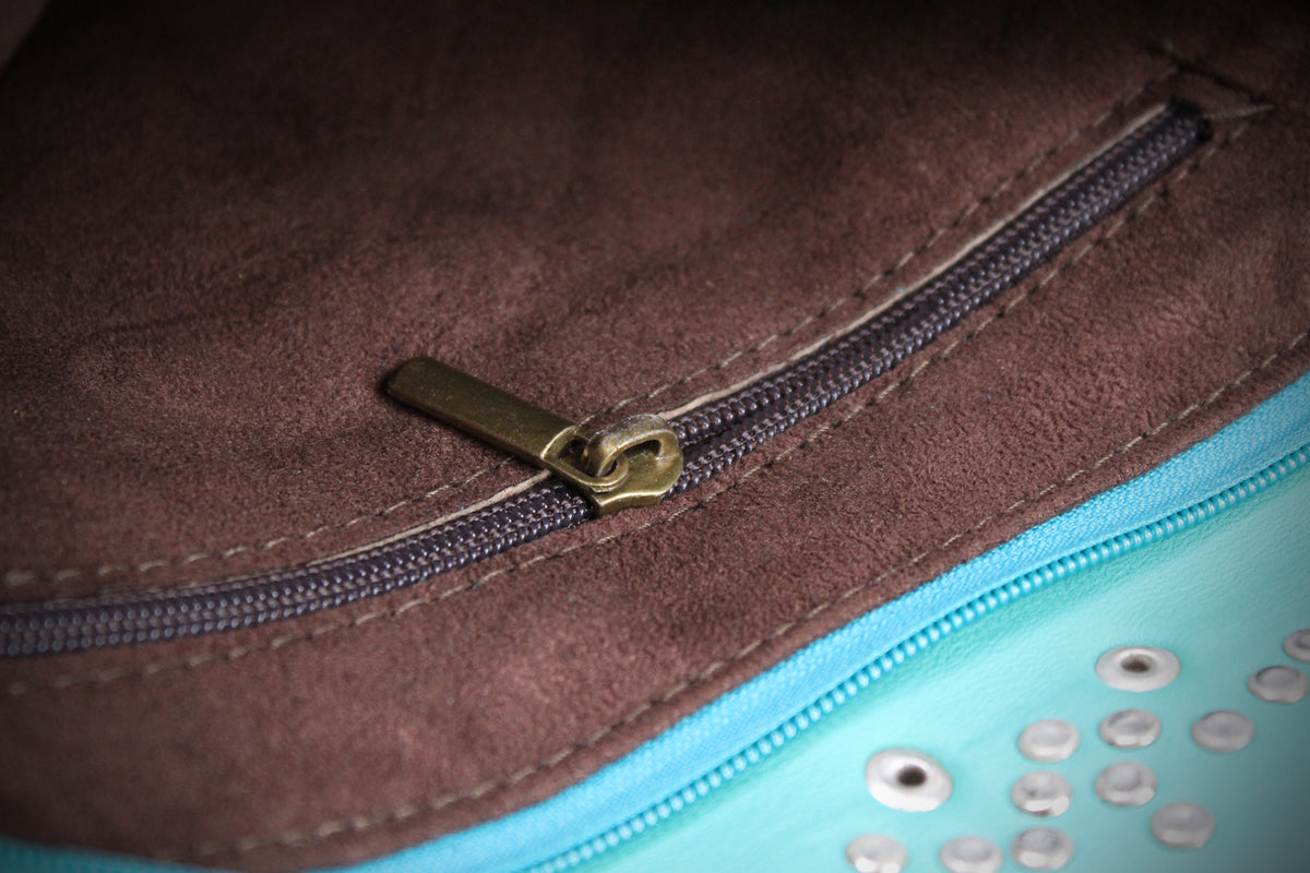 Close-up of a brown suede or faux-suede zipper compartment with a brass-toned zipper pull, next to a bright turquoise fabric section with metallic studs.