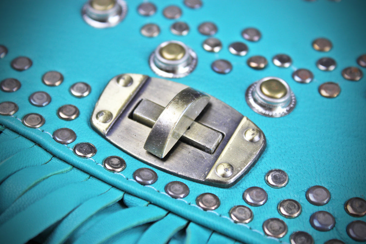 Close-up of a turquoise leather fringe accessory with silver metal studs, snaps, and a central silver clasp on a turquoise background.