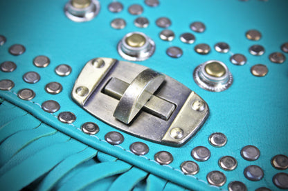 Close-up of a turquoise leather fringe accessory with silver metal studs, snaps, and a central silver clasp on a turquoise background.