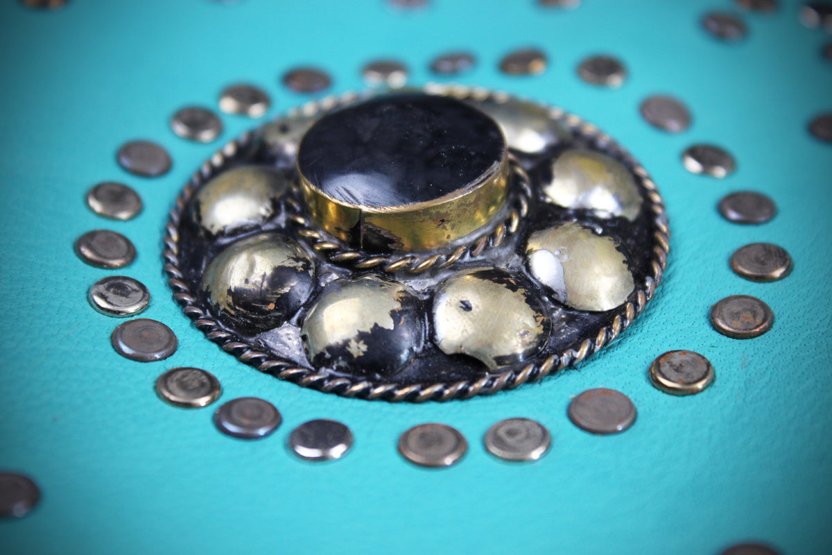 A close-up of a turquoise leather or faux-leather surface with a circular decorative metal stud cluster in the center and a ring of matching studs around it.