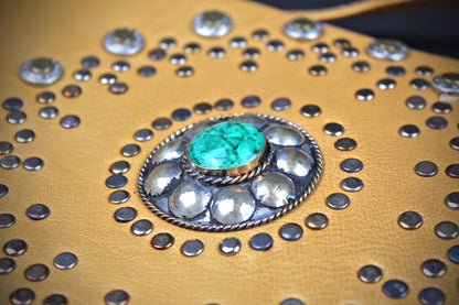 A close-up of a tan leather item heavily decorated with silver-tone studs and a central concho featuring a round turquoise stone.