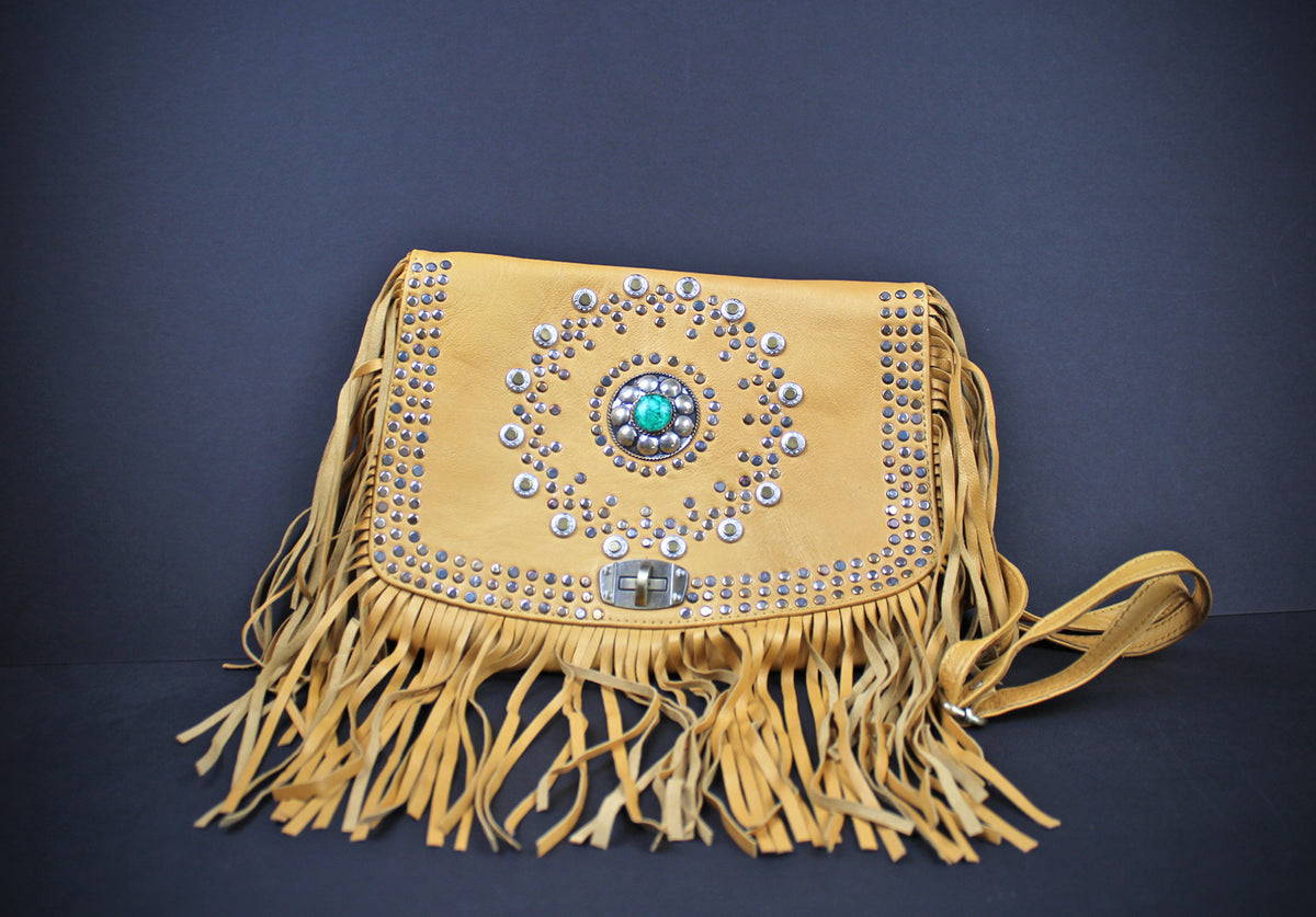 A tan suede fringe bag with silver studs and a turquoise stone centerpiece, displayed on a dark gray background.