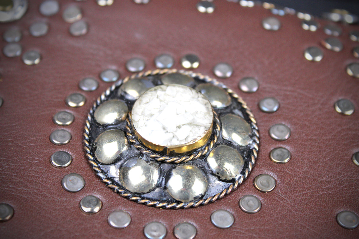 A close-up of a brown leather item decorated with silver-tone studs and a central round metal emblem featuring a white marbled stone.