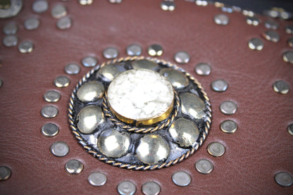 A close-up of a brown leather item decorated with silver-tone studs and a central round metal emblem featuring a white marbled stone.