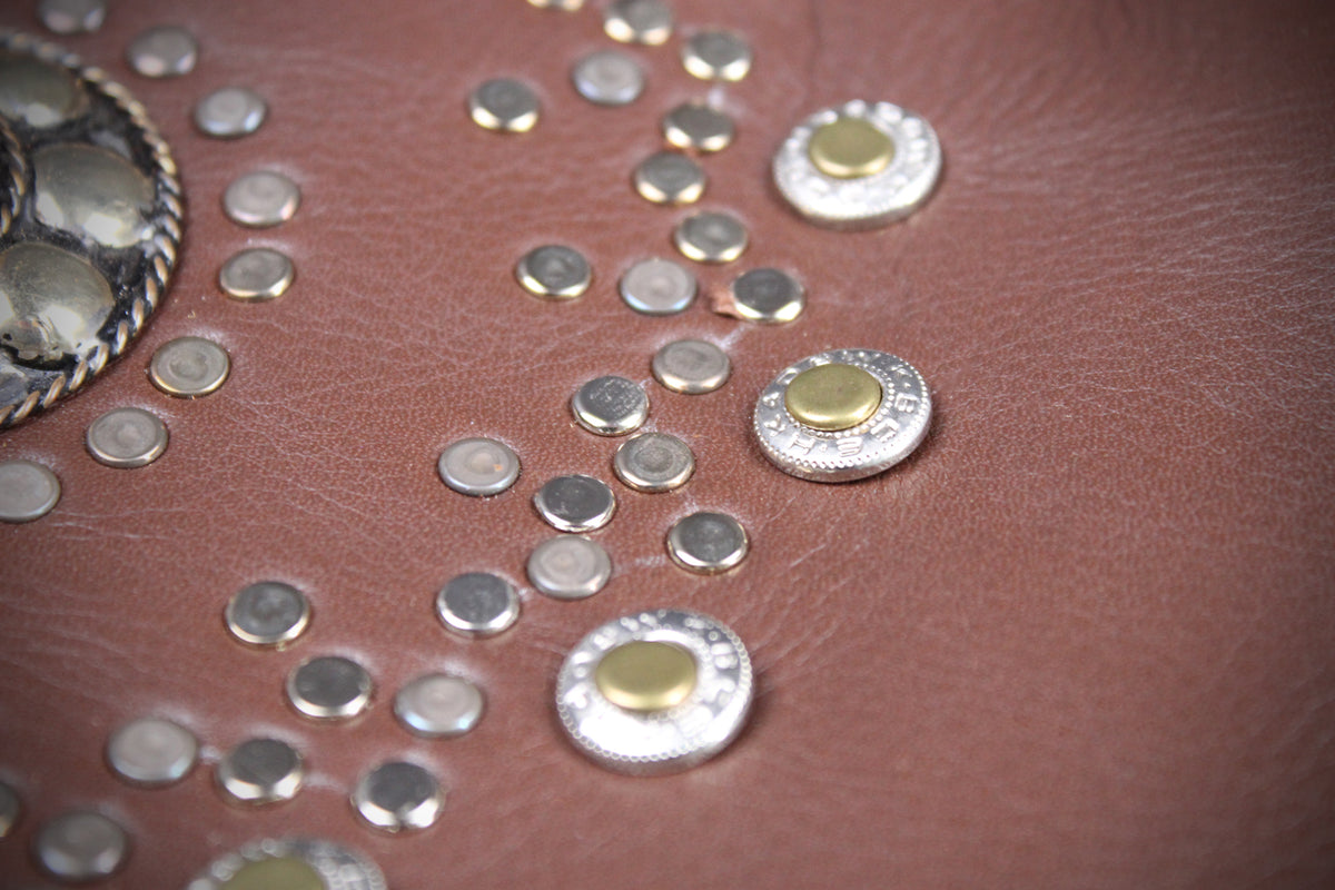 Close-up of a brown leather surface decorated with scattered silver and gold-tone studs and rivets, with part of a metallic embellished circular object visible at the upper left.