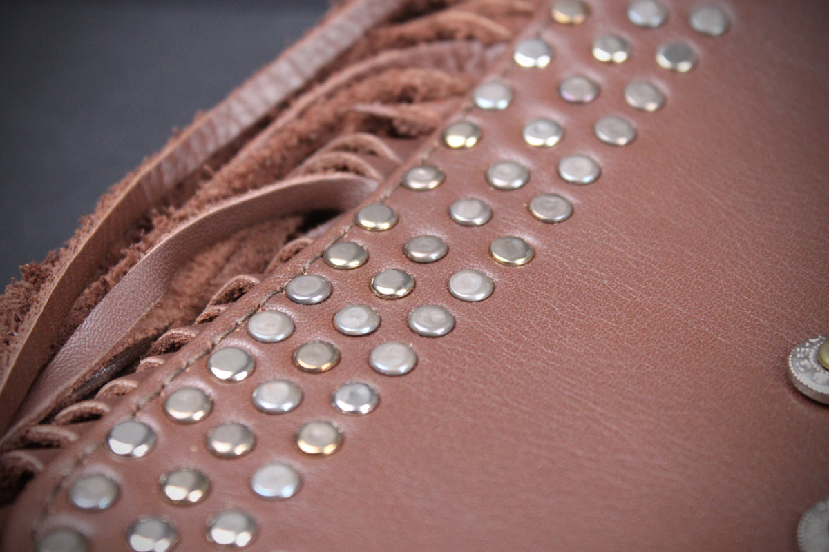 Close-up of a brown leather or faux-leather accessory with fringe, braided trim, and evenly spaced round silver studs.