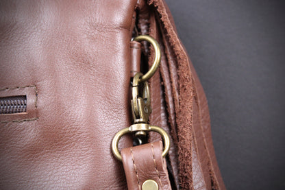Close-up detail of a brown leather bag showing a brass-tone metal swivel clasp, D-ring, and strap attachment against a dark gray background.