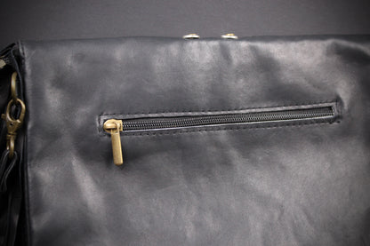 Close-up of a black leather or faux-leather bag with a horizontal zippered pocket and gold-tone zipper pull, shown against a dark background.
