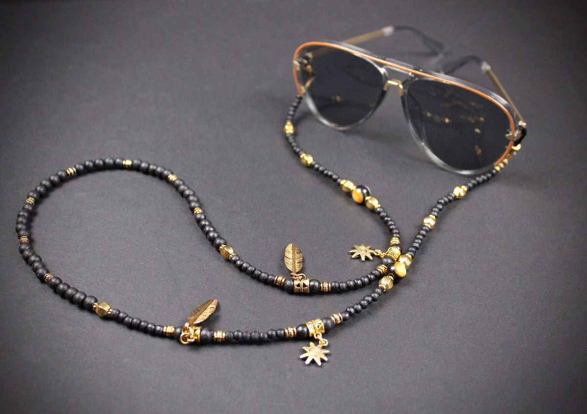 A beaded sunglasses chain strap in black and gold, featuring feather and sunburst charms, shown draped over a pair of aviator sunglasses on a dark gray background.