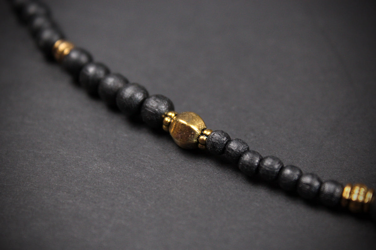 A close-up of a beaded bracelet or necklace made of matte black round beads with small gold spacer accents and a larger central gold bead, photographed on a dark gray background.