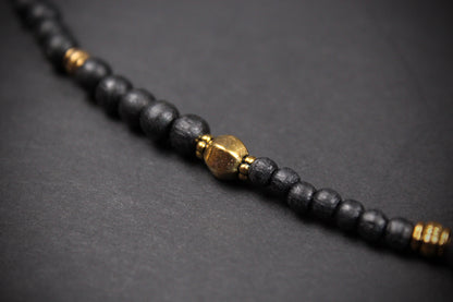 A close-up of a beaded bracelet or necklace made of matte black round beads with small gold spacer accents and a larger central gold bead, photographed on a dark gray background.
