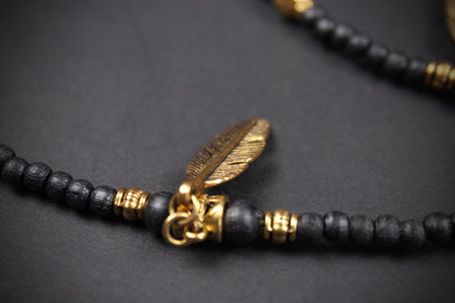 A close-up of a beaded necklace with matte black round beads, small gold spacer beads, and a textured gold feather-shaped pendant on a dark gray background.