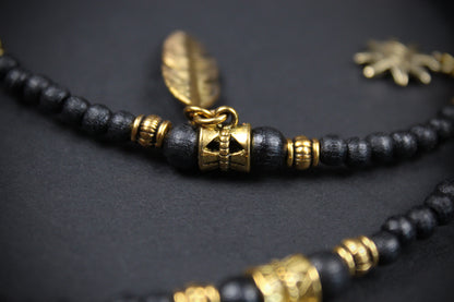 A close-up of a beaded necklace featuring matte black wooden beads, polished gold-toned spacer beads, and a central gold-toned feather charm on a dark textured background.