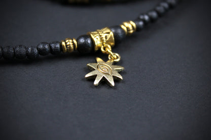 A close-up of a beaded necklace featuring matte black round beads, textured gold spacer beads, and a dangling gold-toned starburst charm with an eye-of-providence symbol in the center, set against a dark gray background.