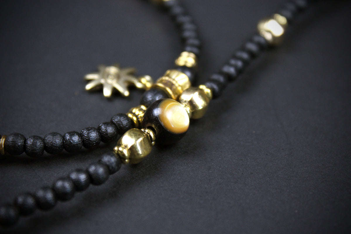 A close-up of a beaded necklace featuring matte black wooden beads, polished gold metal spacers, a round gold tiger's eye bead, and a small gold sun charm, set against a dark textured background.
