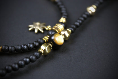 A close-up of a beaded necklace featuring matte black wooden beads, polished gold metal spacers, a round gold tiger's eye bead, and a small gold sun charm, set against a dark textured background.