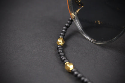 A close-up of a black beaded chain accessory with gold-toned decorative beads, draped over the edge of clear sunglasses on a dark gray surface.