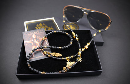 A black beaded necklace with gold-toned charms and spacer beads, displayed in a black presentation box alongside sunglasses, a card, and a small square box on a dark background.