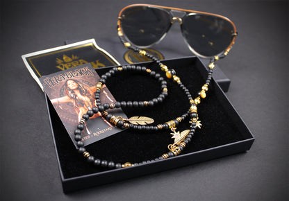 A pair of aviator-style sunglasses with a matching beaded neck strap and gold-toned charms, displayed in a black presentation box with a card on a dark background.