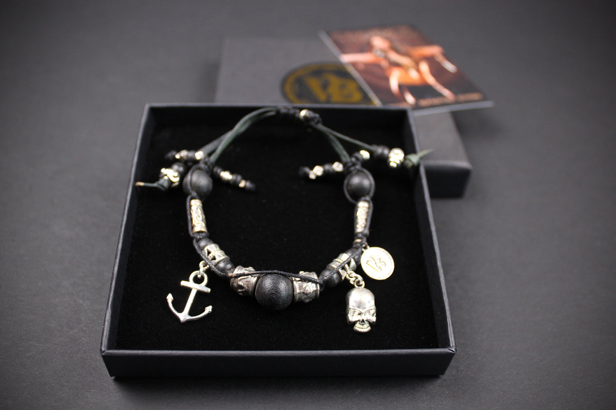 A beaded bracelet with black and metallic silver beads, featuring charms including an anchor, a skull, and a bell, displayed in an open black presentation box.