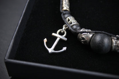 A close-up of a black cord bracelet with a silver anchor charm, skull beads, and a dark round bead, displayed inside a black presentation box.