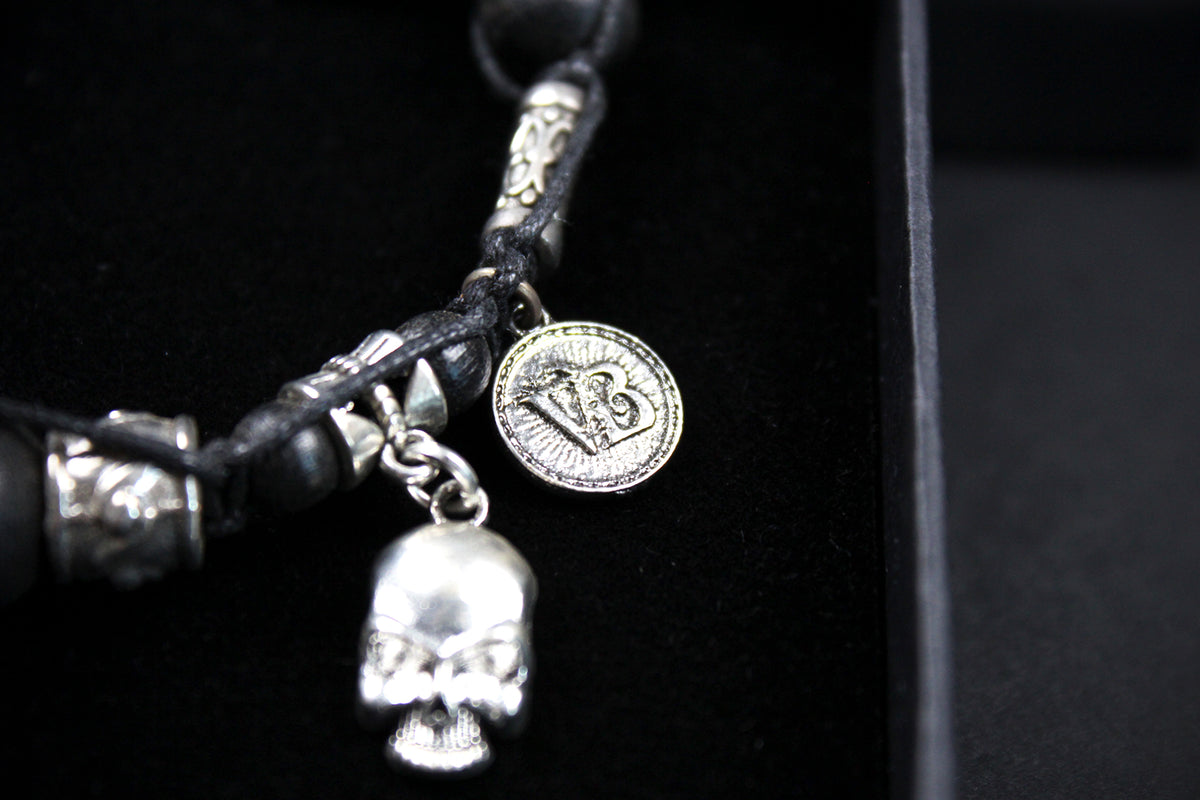 A close-up of a gothic-style black leather bracelet with silver-toned metal accents, including a skull charm, a round medallion charm, and textured silver beads, displayed on a black velvet surface.