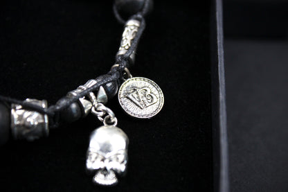 A close-up of a gothic-style black leather bracelet with silver-toned metal accents, including a skull charm, a round medallion charm, and textured silver beads, displayed on a black velvet surface.