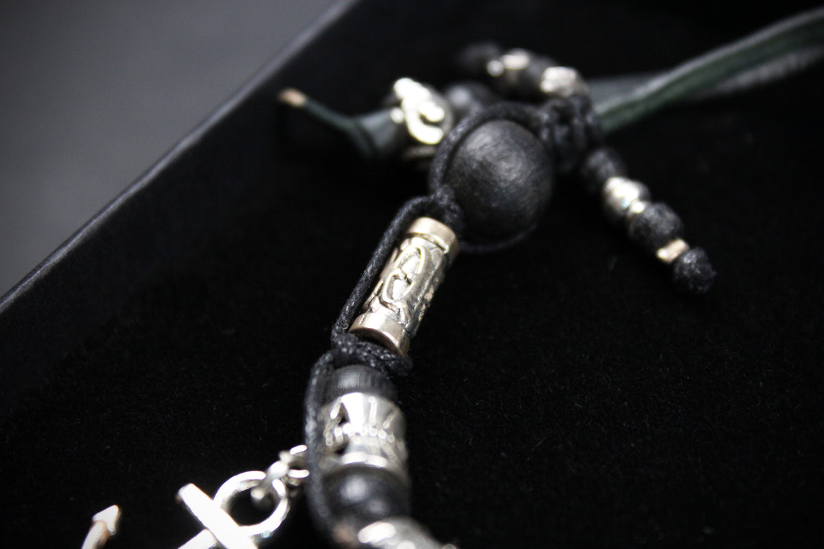 A close-up of a black cord bracelet with black beads, silver-toned metal spacers, and a silver cross charm resting on a black velvet jewelry box.