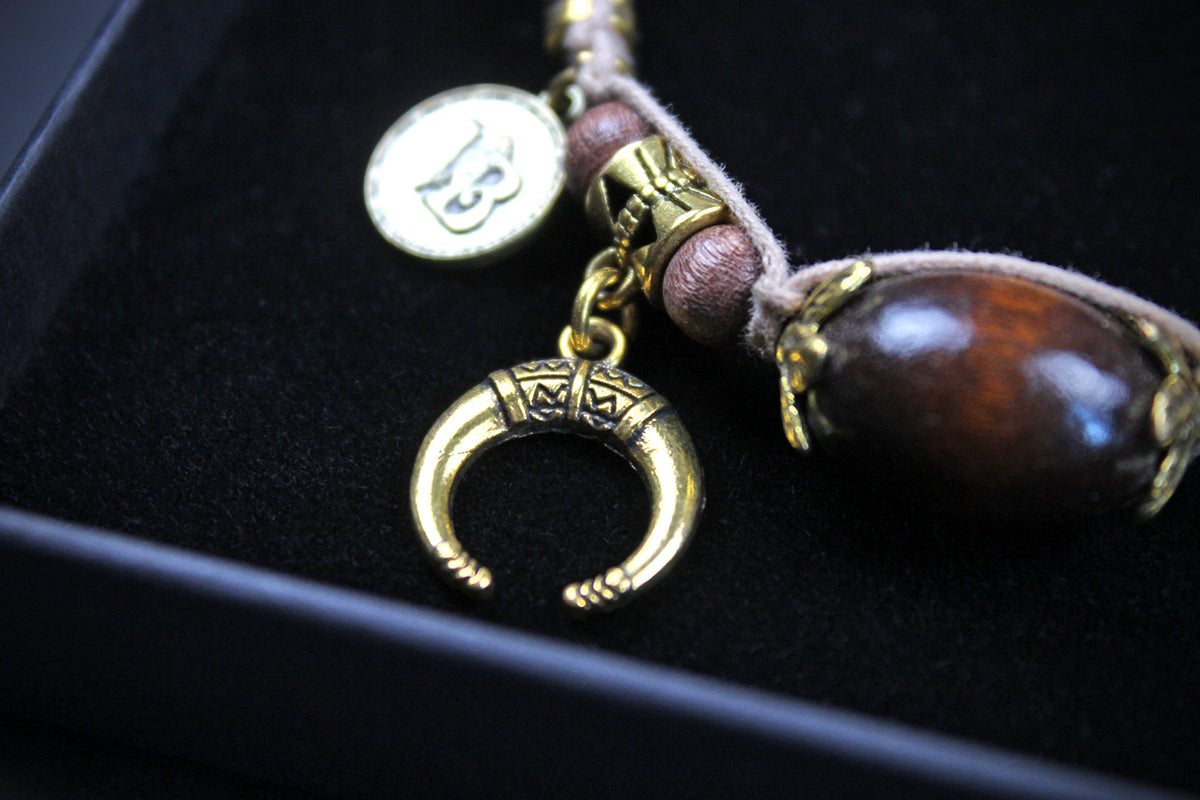 A close-up of a jewelry charm or pendant set displayed in a black box. The set includes a gold-toned crescent moon charm, a round gold-toned coin charm, brown beads, and a large polished dark brown wooden bead with gold-toned caps, all resting on a black velvet surface.