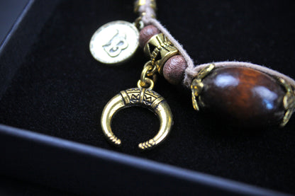 A close-up of a jewelry charm or pendant set displayed in a black box. The set includes a gold-toned crescent moon charm, a round gold-toned coin charm, brown beads, and a large polished dark brown wooden bead with gold-toned caps, all resting on a black velvet surface.